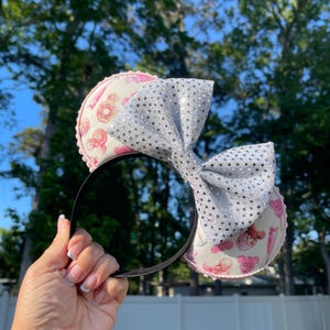 Könnte beinhalten: Ein schwarzes Stirnband mit zwei runden Ohren mit einem rosa-weißen Muster aus Donuts und Eiscreme. Eine große silberne Pailletten-Schleife ist zwischen den Ohren zentriert. Das Stirnband wird vor einem Hintergrund aus Bäumen und blauem Himmel gehalten.