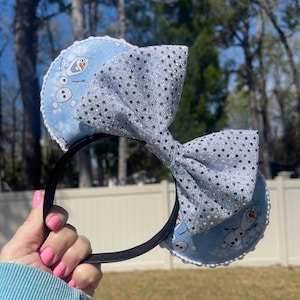 May include: A black headband with blue ears featuring Olaf from Frozen and a large, sparkly silver bow. The ears have a white scalloped trim. The bow is covered in silver sequins. The headband is held by a person.