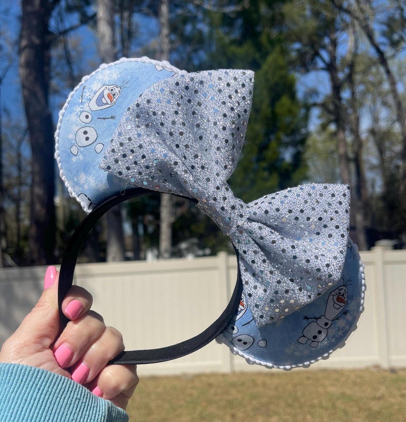 May include: A black headband with blue ears featuring Olaf from Frozen and a large, sparkly silver bow. The ears have a white scalloped trim. The bow is covered in silver sequins. The headband is held by a person.