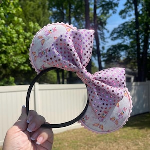 May include: A headband featuring two circular ears with a white fabric base printed with a repeating Daisy Duck pattern. The edges of the ears are trimmed with small pink pom-poms. A large, central bow made of shiny, light pink sequined fabric sits between the ears.