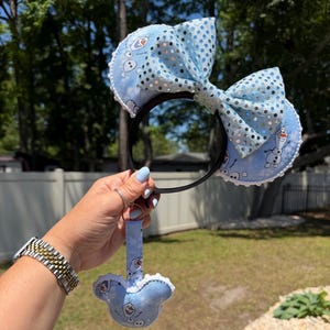 May include: A light blue headband with a large bow and sequin accents. The headband features two round ears with an Olaf character print. A matching Olaf-shaped keychain is attached. The headband is held up by a person.