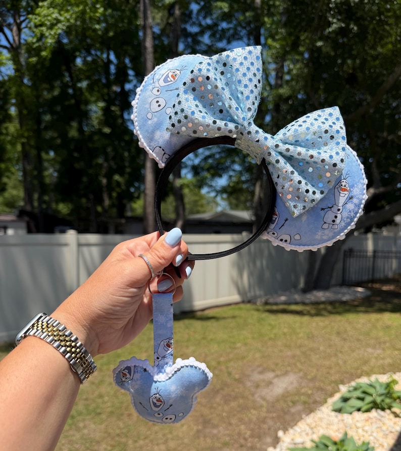 May include: A light blue headband with a large bow and sequin accents. The headband features two round ears with an Olaf character print. A matching Olaf-shaped keychain is attached. The headband is held up by a person.