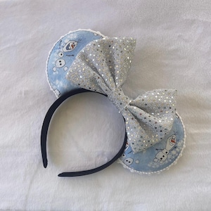 May include: A black headband with two round blue ears featuring Olaf from Frozen. A large silver sequined bow sits between the ears. The ears have a white lace trim.