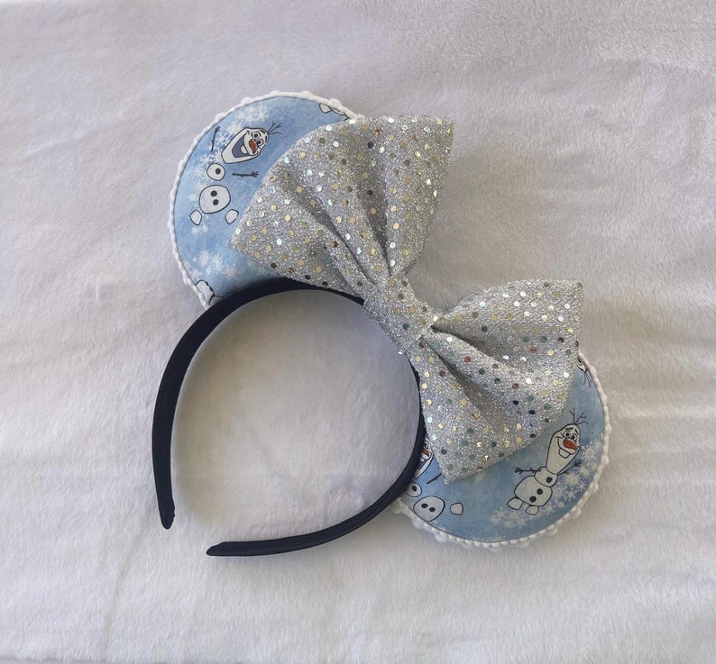 May include: A black headband with two round blue ears featuring Olaf from Frozen. A large silver sequined bow sits between the ears. The ears have a white lace trim.