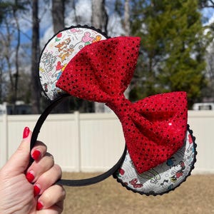 May include: Black headband with two round ears featuring a cartoon design and a large red sequined bow. The ears have a white background with colorful cartoon characters and a black scalloped trim. The bow is a vibrant red with black sequins.
