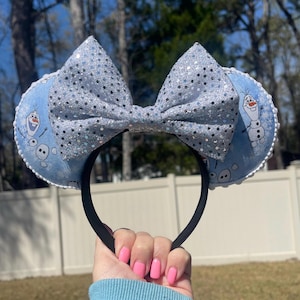May include: A pair of mouse ears featuring a light blue design with Olaf from Frozen. The ears have a large, sparkly silver bow and a black headband. The ears are trimmed with white lace.