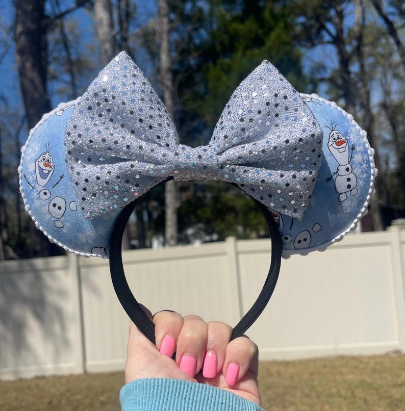 May include: A pair of mouse ears featuring a light blue design with Olaf from Frozen. The ears have a large, sparkly silver bow and a black headband. The ears are trimmed with white lace.
