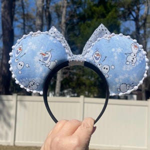 May include: Blue mouse ears featuring Olaf from Frozen, with a snowflake pattern. The ears are trimmed with white pom-poms and have a silver glitter bow. The headband is black.
