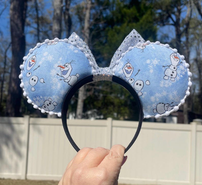 May include: Blue mouse ears featuring Olaf from Frozen, with a snowflake pattern. The ears are trimmed with white pom-poms and have a silver glitter bow. The headband is black.