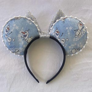 May include: A headband featuring two round, light blue ears with a pattern of Olaf the snowman and snowflakes. The ears are trimmed with white pom-poms and have a silver glitter bow. The headband itself is black.