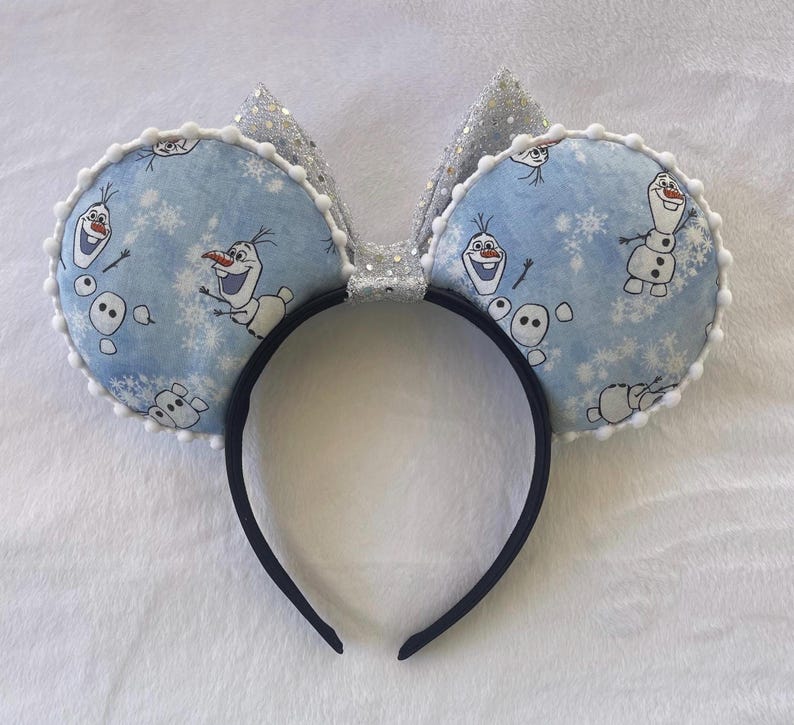 May include: A headband featuring two round, light blue ears with a pattern of Olaf the snowman and snowflakes. The ears are trimmed with white pom-poms and have a silver glitter bow. The headband itself is black.