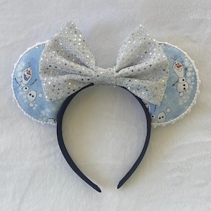 May include: A pair of mouse ears featuring a large, silver glitter bow and two round ears. The ears have a light blue background with a pattern of Olaf from Frozen. The headband is black.