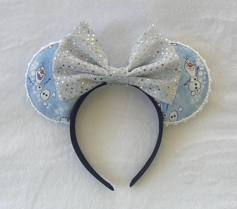 May include: A pair of mouse ears featuring a large, silver glitter bow and two round ears. The ears have a light blue background with a pattern of Olaf from Frozen. The headband is black.