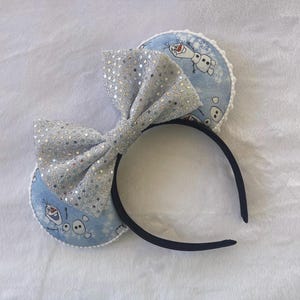 May include: A black headband with two light blue circular ear accents featuring a cartoon snowman design. A large, silver sequined bow is centered on the headband. The ears have a white trim.