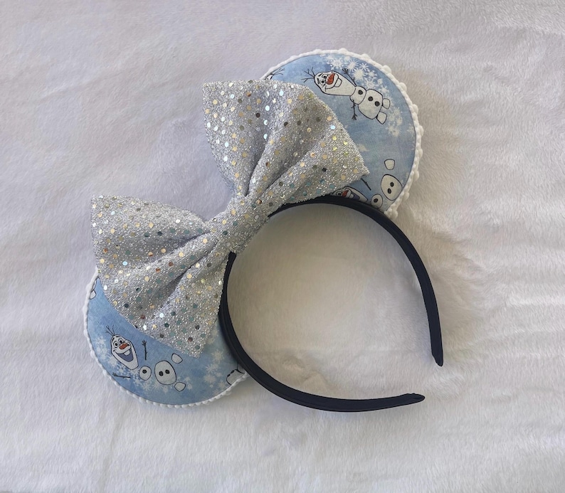 May include: A black headband with two light blue circular ear accents featuring a cartoon snowman design. A large, silver sequined bow is centered on the headband. The ears have a white trim.