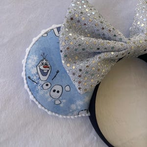May include: A pair of mouse ears featuring a light blue ear with a print of Olaf from Frozen and snowflakes. The ears have a white trim and a large silver sequined bow. The headband is black.