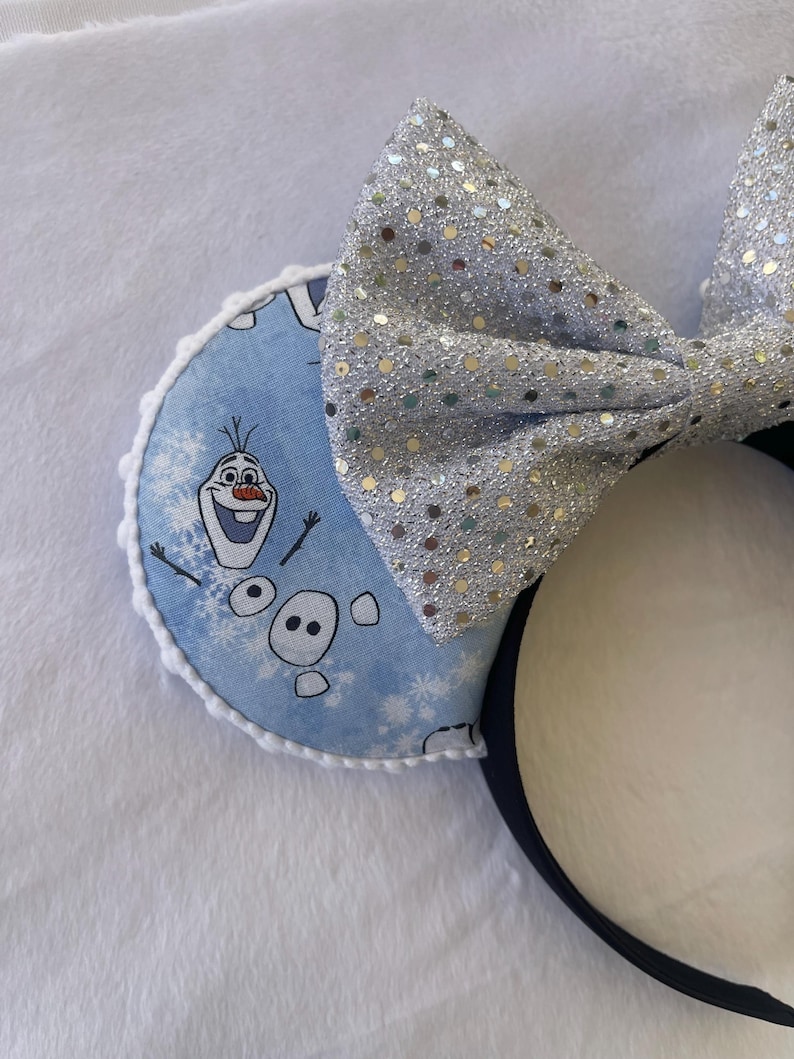 May include: A pair of mouse ears featuring a light blue ear with a print of Olaf from Frozen and snowflakes. The ears have a white trim and a large silver sequined bow. The headband is black.