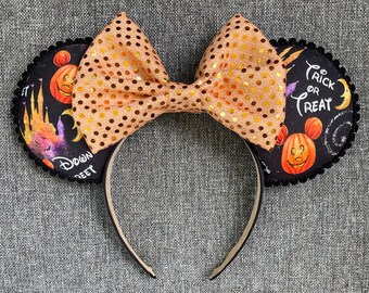 May include: A headband featuring two round ears with a black fabric base printed with orange pumpkins, a castle, and the text Trick or Treat. A large, shimmering gold polka-dot bow sits between the ears, which are edged with small black pom-pom trim.