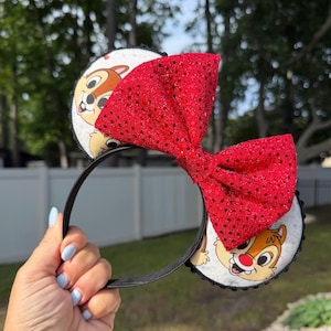 May include: Black headband with Chip and Dale character ears and a large, red sequined bow. The ears feature a white background with the cartoon characters. The bow is made of red sequins.