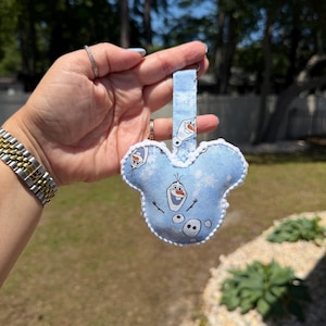 May include: A light blue Mickey Mouse-shaped keychain featuring Olaf from Frozen. The keychain has a white trim and a matching blue strap. The design includes snowflakes and Olaf's face. The background is a blurred outdoor scene.