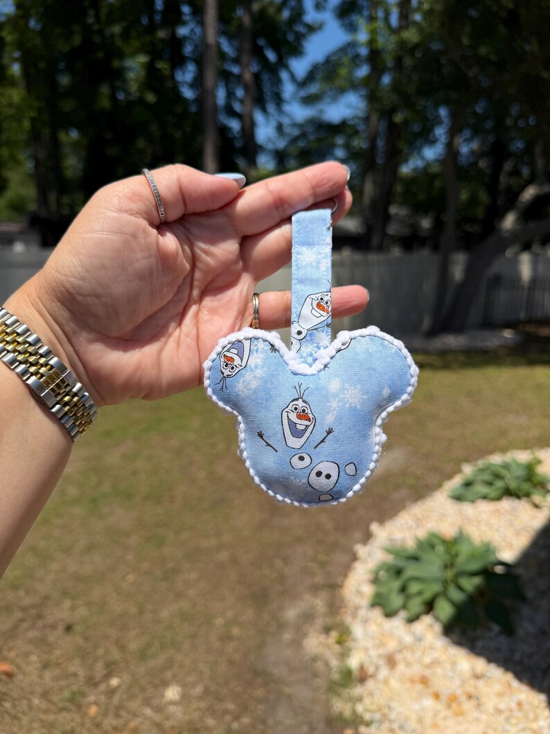 May include: A light blue Mickey Mouse-shaped keychain featuring Olaf from Frozen. The keychain has a white trim and a matching blue strap. The design includes snowflakes and Olaf's face. The background is a blurred outdoor scene.
