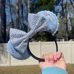 May include: A black headband with light blue ears featuring a cartoon Olaf design. The ears are trimmed with white lace and a large, sparkly silver bow. The bow and ears are decorated with silver glitter.