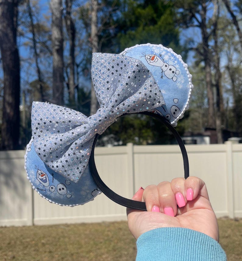 May include: A black headband with light blue ears featuring a cartoon Olaf design. The ears are trimmed with white lace and a large, sparkly silver bow. The bow and ears are decorated with silver glitter.