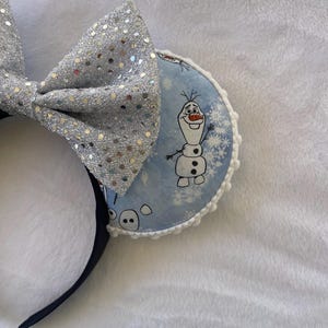 May include: A pair of mouse ears featuring a silver sequined bow and round ear pieces. The ear pieces have a light blue background with a cartoon image of Olaf the snowman and snowflakes. The headband is black.