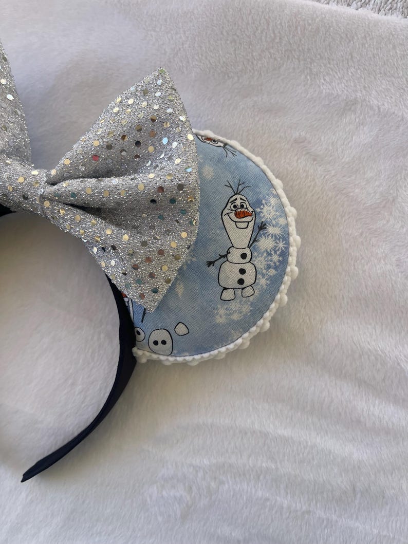 May include: A pair of mouse ears featuring a silver sequined bow and round ear pieces. The ear pieces have a light blue background with a cartoon image of Olaf the snowman and snowflakes. The headband is black.