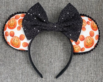 May include: A headband featuring two circular ears with a white background and an orange pumpkin pattern. Each pumpkin is shaped like a mouse head with a carved face. A large, black, glittery fabric bow sits between the ears, and the edges of the ears are trimmed with small black pom-poms.