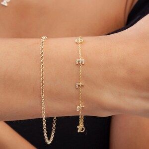 May include: Two gold bracelets on a wrist. One bracelet is a simple chain, the other has small letters with diamonds on it.