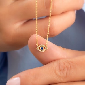 May include: A gold chain necklace with a small evil eye pendant. The pendant is surrounded by small clear stones and has a blue center.
