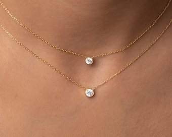 May include: Two gold necklaces with a small round clear stone pendant on each. The necklaces are layered on a person's neck.