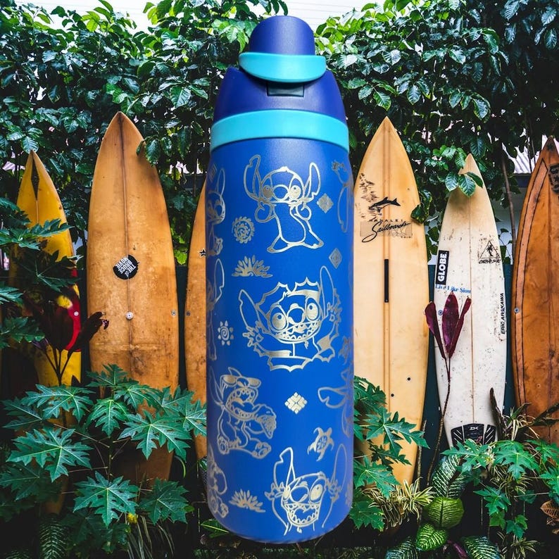 May include: A blue water bottle with a silver Stitch character design. The bottle has a light blue lid and accents. The background features surfboards and green foliage, evoking a tropical atmosphere.