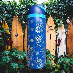 May include: A blue water bottle with a light blue band and lid. The bottle is decorated with gold-coloured cartoon character designs. Surfboards are in the background, with green foliage in the foreground.
