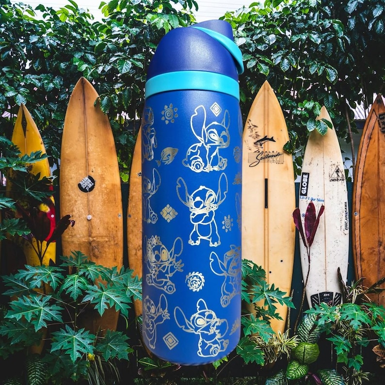 May include: A blue water bottle with a light blue band and lid. The bottle is decorated with gold-coloured cartoon character designs. Surfboards are in the background, with green foliage in the foreground.