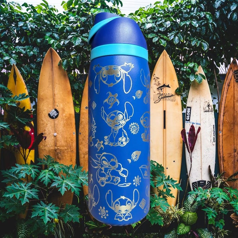 May include: A blue water bottle with a gold character design. The bottle has a blue lid and a light blue band. The background features surfboards and green foliage. The bottle is approximately 25 cm tall.