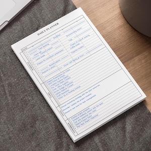 May include: A white daily planner notepad with a detailed layout for scheduling tasks. The notepad is on a dark grey surface next to a laptop and a potted plant. The notepad has the words "DAILY PLANNER" at the top.