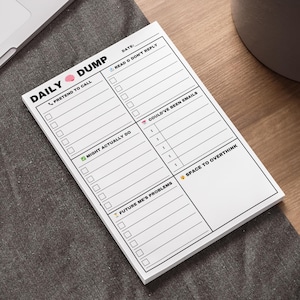 Peut inclure: Un bloc-notes blanc avec le texte "DAILY DUMP" et diverses cases à cocher et invites. Le bloc-notes est sur une surface gris foncé à côté d'un ordinateur portable et d'un stylo. Le bloc-notes est conçu pour la gestion quotidienne des tâches.