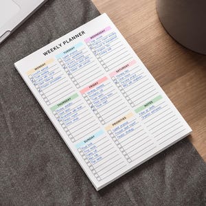 Puede incluir: Un bloc de notas de planificación semanal blanco con un encabezado colorido para cada día de la semana. El bloc de notas tiene casillas de verificación para tareas y una sección para notas y prioridades. Un portátil, un bolígrafo y una planta están en el fondo.