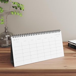 May include: A white desk calendar for May 2026 with a black spiral binding. The calendar is on a wooden surface, next to a glass vase with a plant and a stack of magazines.