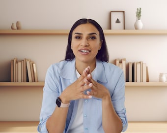 May include: A woman with long dark hair is wearing a light blue button-down shirt. She is sitting in front of a bookshelf with books and decorative items.