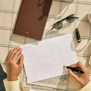 Person writing on an August 2027 printable monthly calendar at a desk with stationery and a notebook.