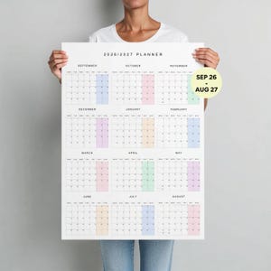 Pode incluir: Um calendário planejador 2026-2027 apresentando doze grades mensais de setembro a agosto. Cada mês exibe os dias da semana com colunas específicas destacadas em cores pastel suaves. Um emblema circular amarelo diz SEP 26 - AUG 27.