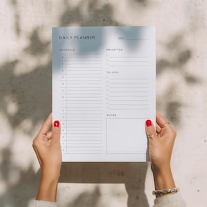 Hand holding a printed daily planner page against a neutral grey wall, showing the minimalist black-and-white layout with lined schedule, notes box, and task checklist areas.
