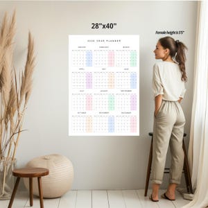 May include: A 28" x 40" 2026 year planner calendar with pastel color-coded months. The calendar is hanging on a white wall. A woman stands next to the calendar. The calendar is a wall decor item.