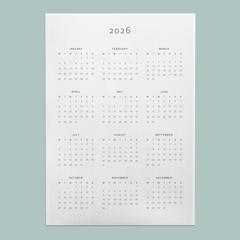 A printed 2026 year at a glance calendar on white A4 paper with a minimalist design in grey modern font, laid flat on a neutral wooden desk with soft natural lighting.