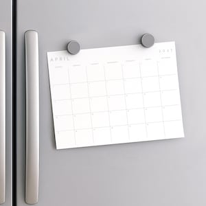 Printable 2027 monthly calendar attached to a wall with magnets beside a modern silver radiator.