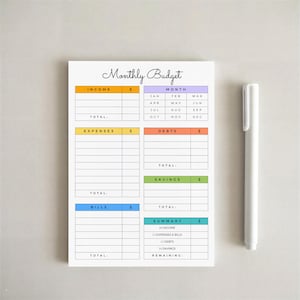 May include: A white notepad with the words "Monthly Budget" in cursive script. The notepad has sections for income, expenses, debts, bills, savings, and a summary. A white pen rests on the right side of the notepad.