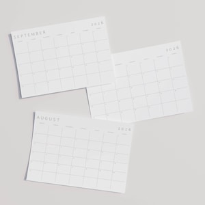 Close-up of a landscape 2026 monthly planner on A4 paper, clean grey lines and modern sans-serif typography, held above a neutral flat surface.